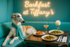 Elegant borzoi in turquoise scarf at turquoise banquette with pretzel, mini pizzas, cocktail & single white flower under “Barkfast at Tiffany’s” neon sign