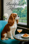 Cavalier King Charles Spaniel on a turquoise chair by a window with a croissant and peach cocktail on a metal table beneath Barkfast at Tiffany’s lettering