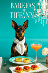 Black and tan Miniature Pinscher wearing white neckerchief seated at metal table with two mini spinach quiches tomato bruschetta crostini a peach cocktail and white dahlia under a crystal chandelier and Barkfast at Tiffany’s sign