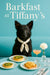Curly black Mudi wearing an ivory scarf seated at a turquoise café table with two spinach quiches apple-topped toast a peach cocktail and a white dahlia under Barkfast at Tiffany’s sign