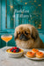 Regal Pekingese seated on a teal banquette beside a marble table with custard topped by raspberries and blueberries three smoked salmon tartlets and a pink cocktail under starlight mirror