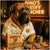 Mastiff wearing a gold chain and custom shirt, seated at a table with a box of deli treats, dollar bills, and snack platters under the neon sign "Dino's Mastiff Muncher".