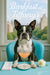 Tuxedo-masked Boston Terrier perched on a turquoise banquette by a city-view window with a blush-hued cocktail, white napkin, and single white flower under “Barkfast at Tiffany’s” script