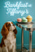 Cavalier King Charles Spaniel looking up at pastel macarons and a cupcake on a metal table under a neon Barkfast at Tiffany’s sign against a teal background