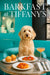 Cream-colored labradoodle seated at turquoise café table with golden muffins tomato-topped toast cheese biscuit and pink cocktail under Barkfast at Tiffanys sign