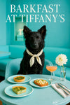 Black Mudi wearing an ivory scarf seated at a turquoise café table with spinach quiches, apple-topped toast, peach cocktail, and white dahlia under Barkfast at Tiffany’s sign