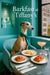 Whippet wearing a teal scarf seated in a turquoise booth with gift boxes and a crystal dog figurine in a display niche next to a table holding lemon tartlets tomato toast a peach cocktail and a glass of water