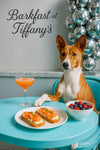 Basenji dog at a Tiffany-blue café table with smoked-salmon toast garnished with chives, a bowl of raspberries and blueberries, and a peach cocktail beside a silver bubble sculpture