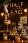 Four squirrels enjoying cashews and almond butter cocktails at a tree-trunk bar under a “Half in the Bag” sign